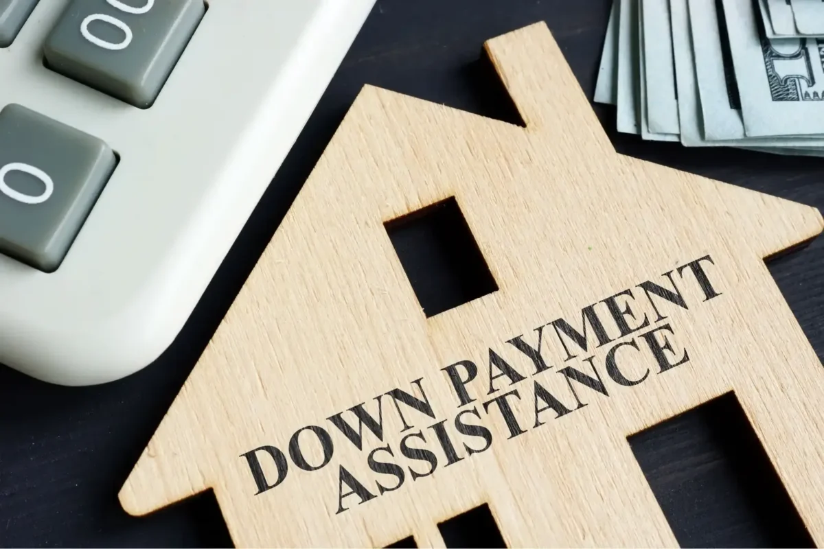 Wooden house cutout reading Down Payment Assistance beside a calculator and cash