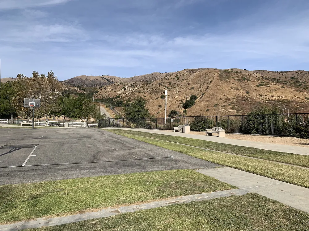 A paved basketball court and lawn at Holleigh Bernson Memorial Park with dry, brush-covered hills rising behind it in Porter Ranch