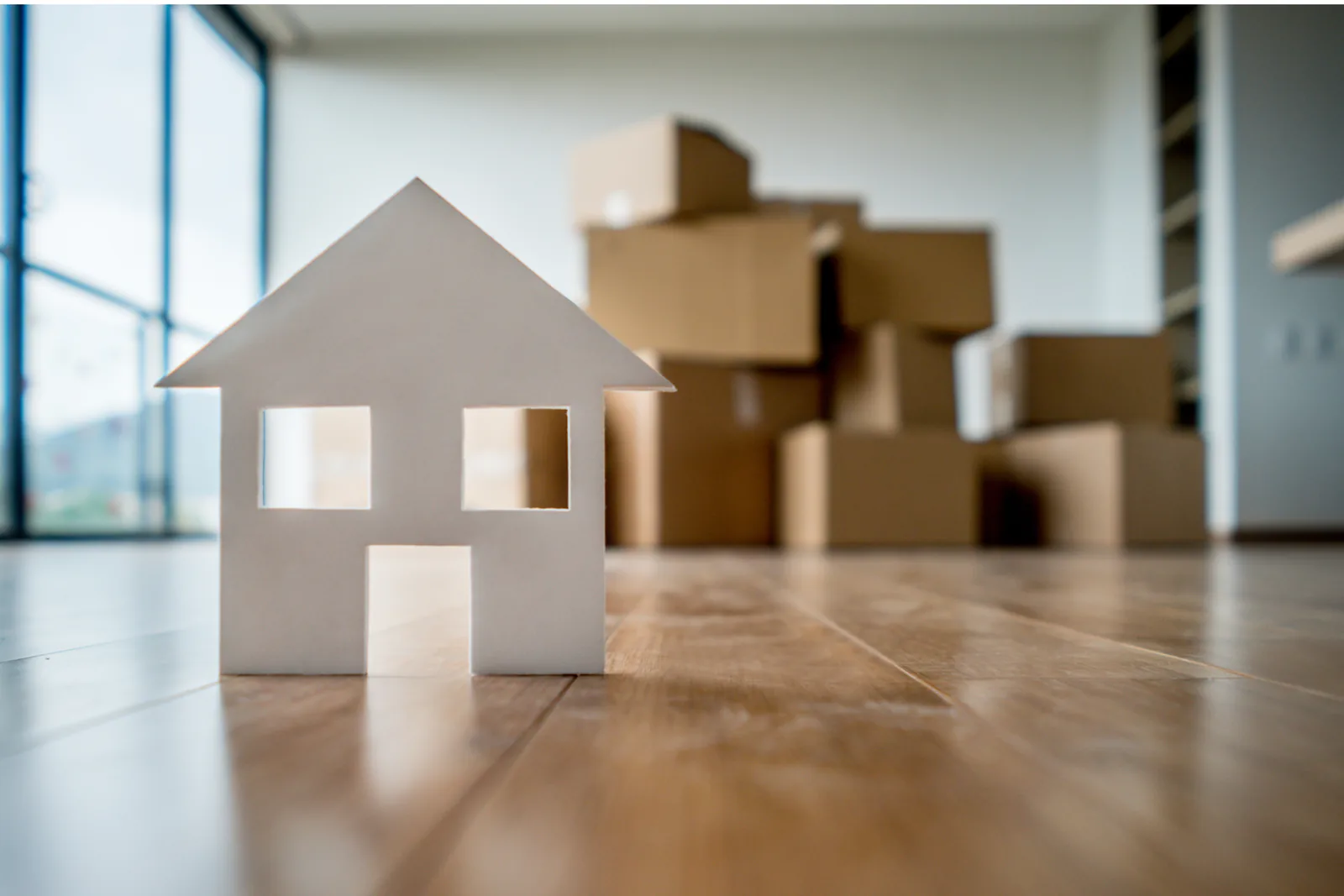 Paper cutout of a house standing on a wood floor in front of stacked moving boxes