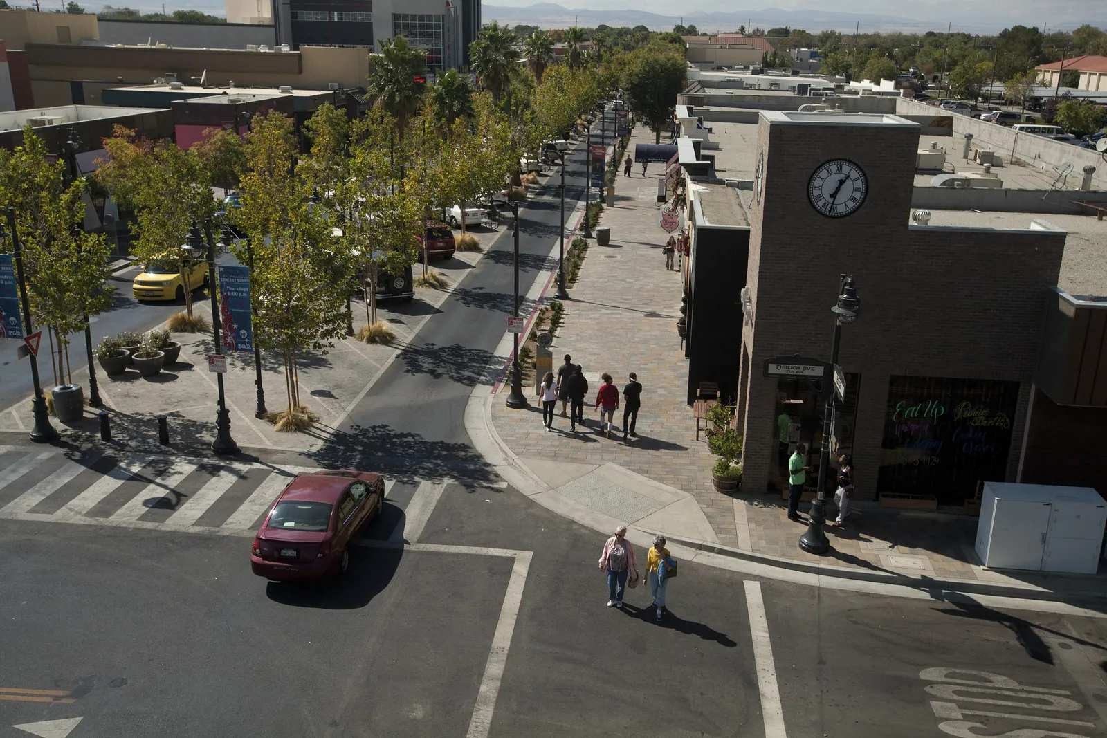 Pedestrians crossing tree-lined Lancaster Boulevard downtown, with shops, a clock tower and wide sidewalks on The BLVD