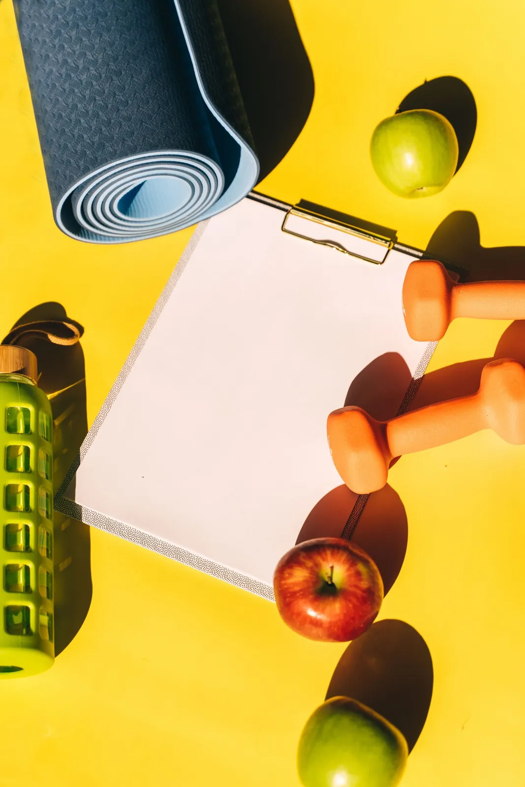 Clipboard, yoga mat, dumbbells and fruit on a yellow background