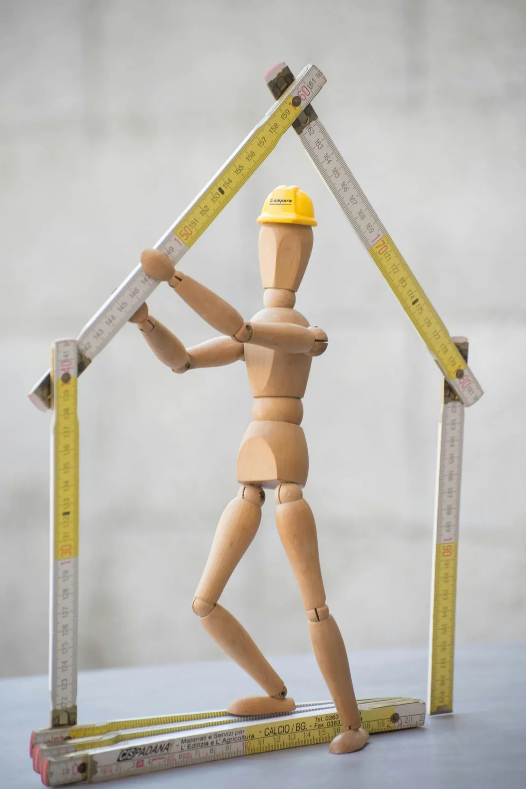 Wooden figure in a yellow hard hat holding folding rulers shaped like a house frame