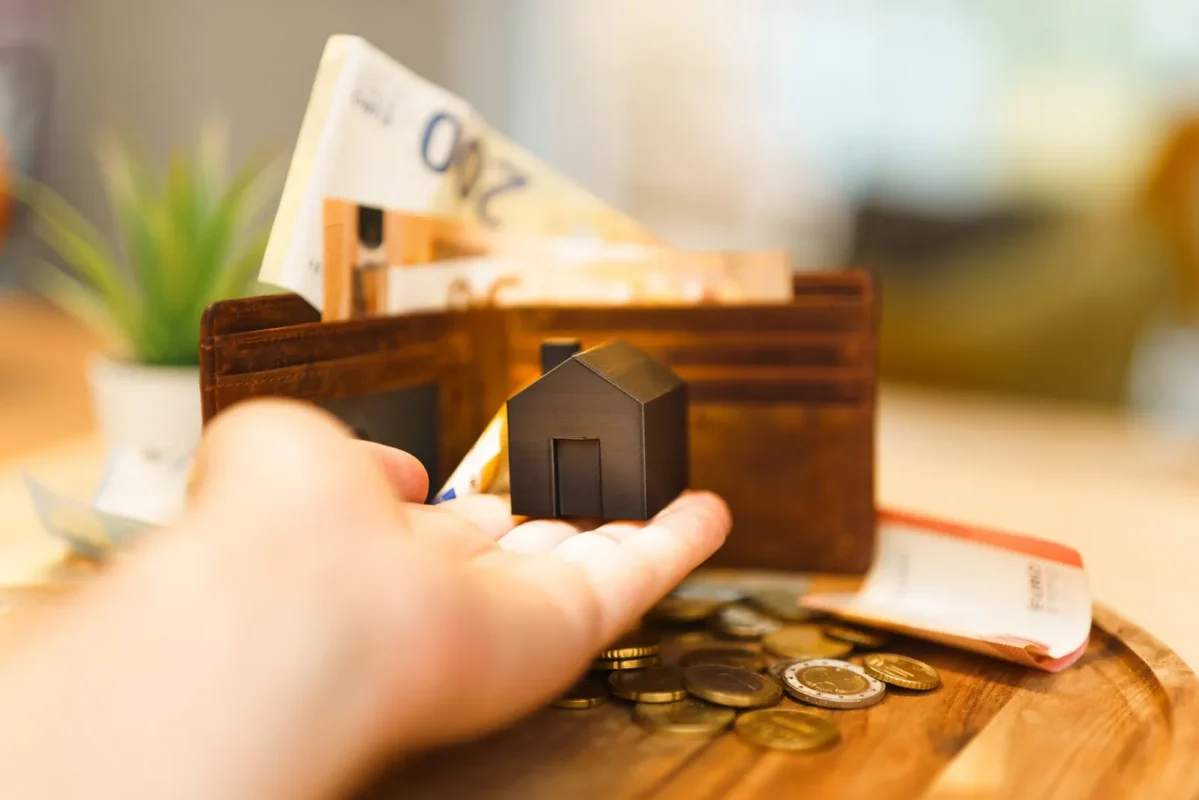 Hand holding a small model house beside a wallet with cash
