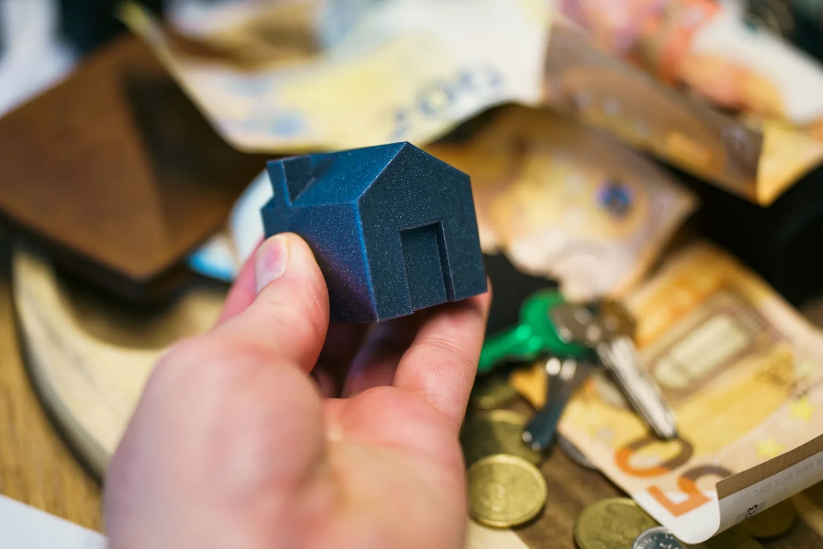 Hand holding a small blue model house over cash and coins