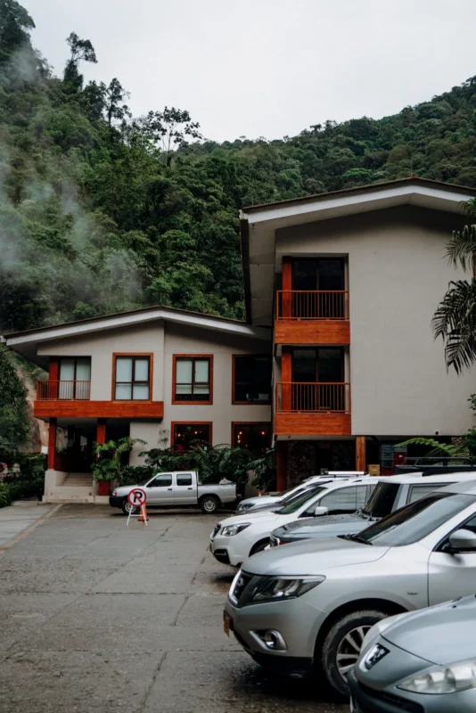 Two-story building with orange balconies and parked cars below wooded hills