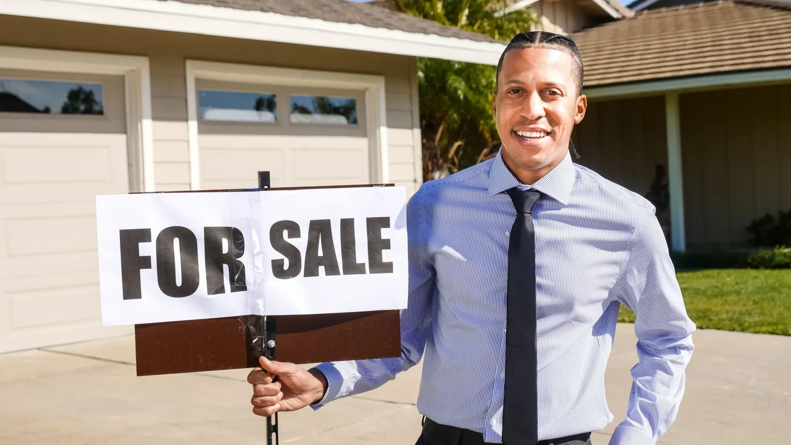 Smiling agent holding a 'For Sale' sign in front of a house