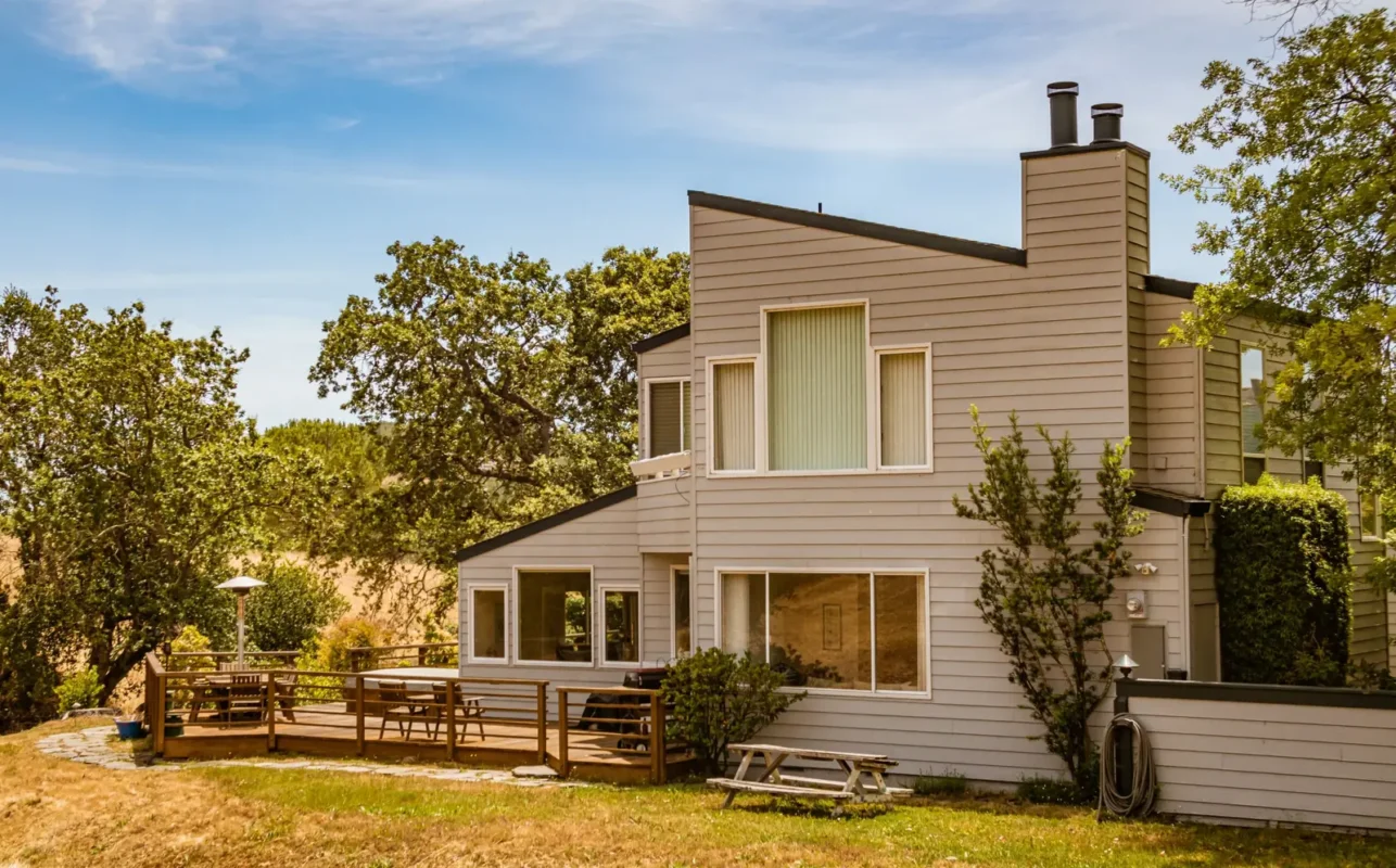 Two-story wood-sided house with a deck and picnic table in a sunny yard