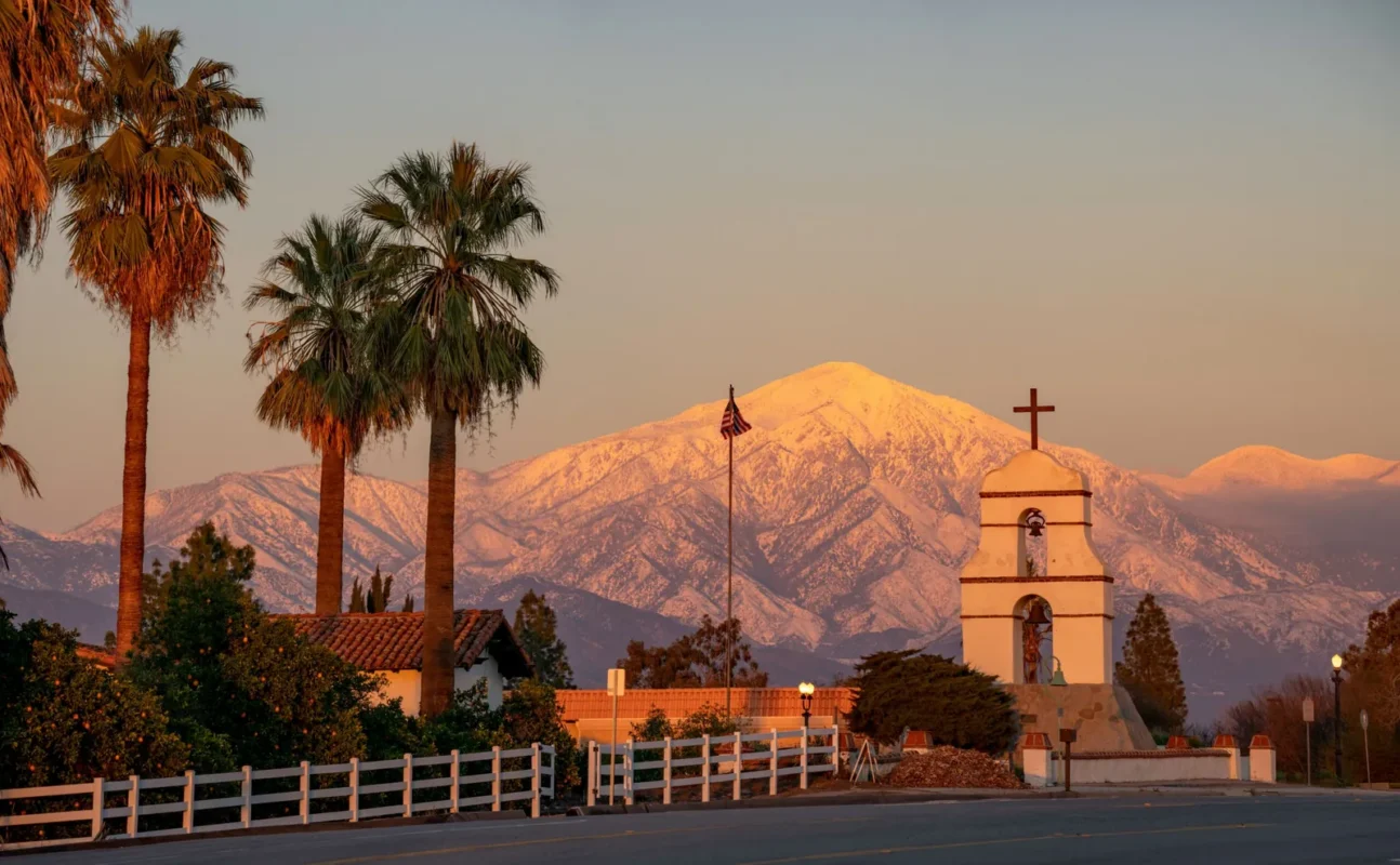 Mission-style bell tower and palm trees before snow-capped mountains at sunset