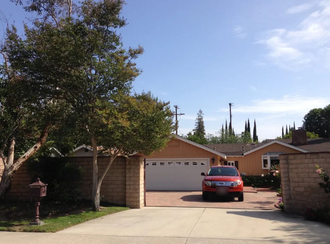 Mature trees shading a brick-edged driveway that leads to a tan single-story ranch house with a two-car garage in Northridge