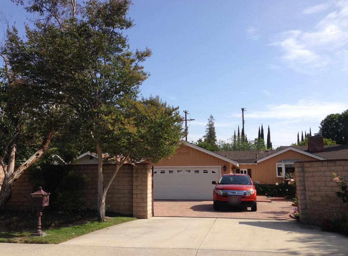 Mature trees shading a brick-edged driveway that leads to a tan single-story ranch house with a two-car garage in Northridge