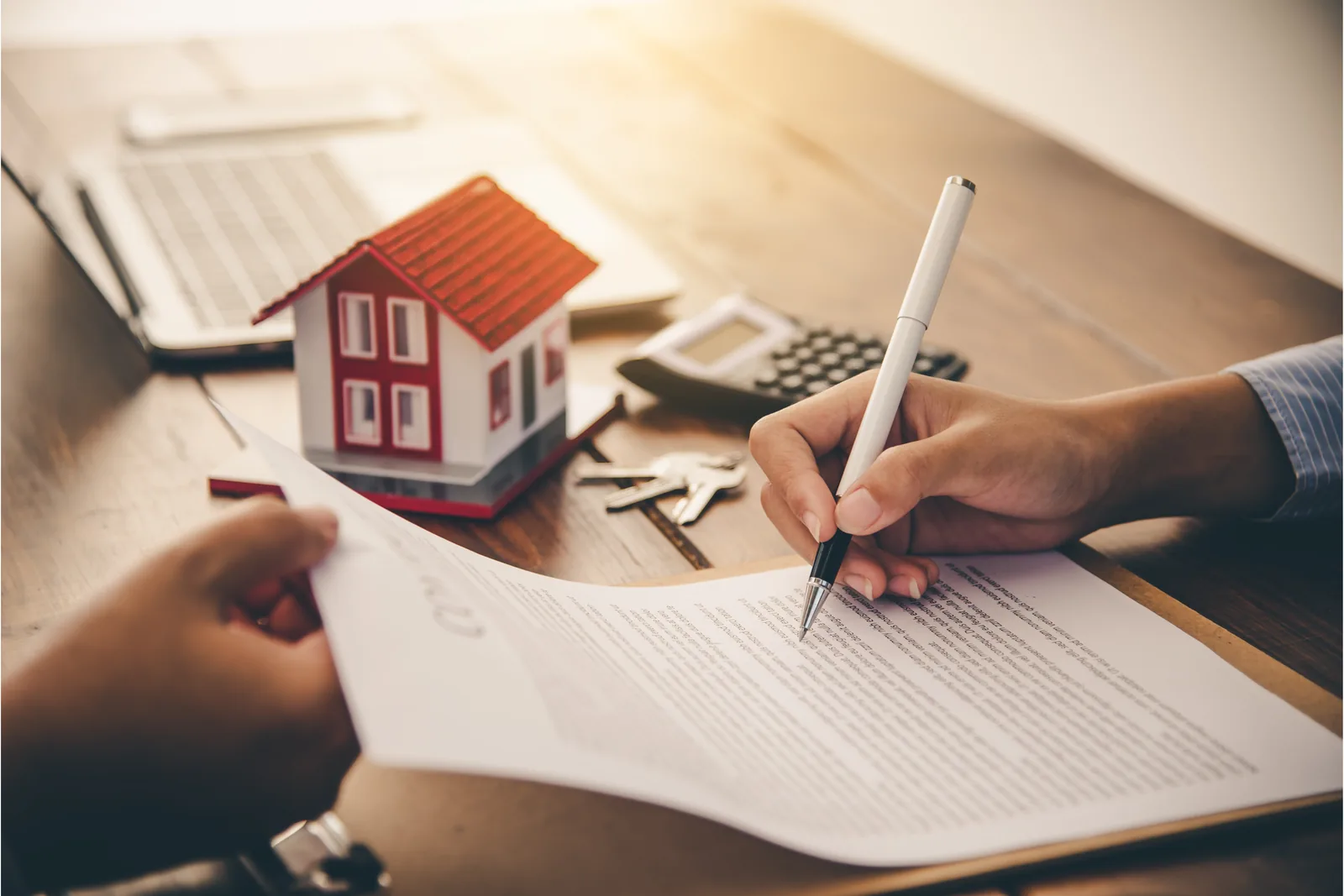 Person signing paperwork beside a small model house, keys and a calculator