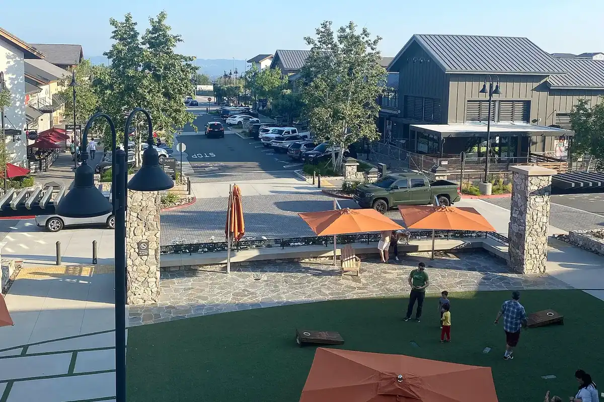 Outdoor plaza at The Vineyards at Porter Ranch with orange umbrellas, a lawn with cornhole boards, stone pillars and modern farmhouse-style shop buildings